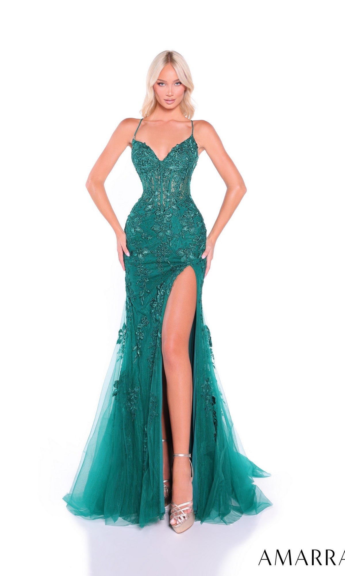 A woman poses against a white background wearing the Amarra 89022, an embroidered-lace long prom dress featuring emerald green fabric, a fitted silhouette, open back, floral details, thin straps, and a high thigh slit.